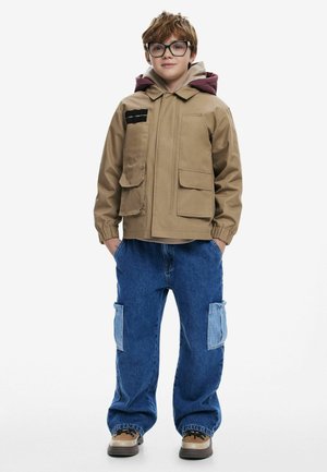 Tans zip-up jack met twee voorzakken en een rode capuchon; gecombineerd met blauwe baggy jeans met twee paspelzakken. Bruine schoenen met zwarte veters.