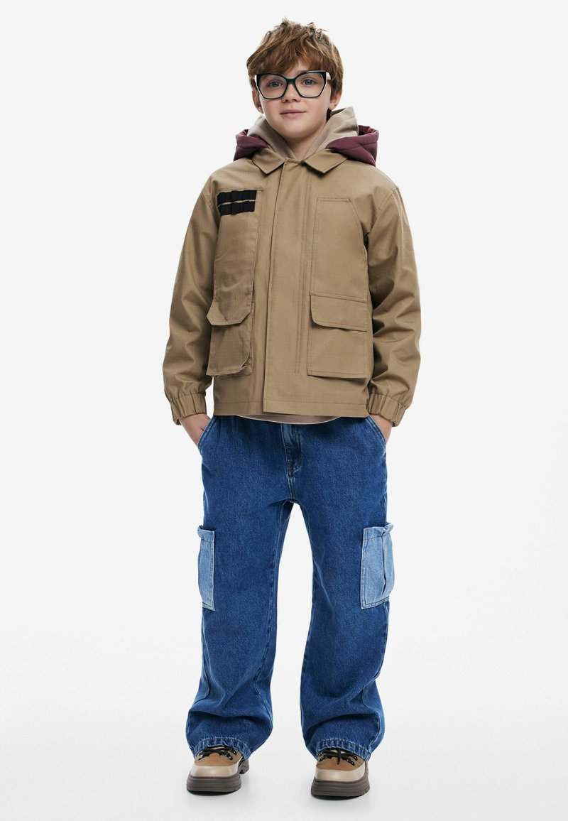 Tan zip-up jacka med två framfickor och en röd huva; matchad med blå baggy jeans med två lagda fickor. Bruna skor med svarta snören.