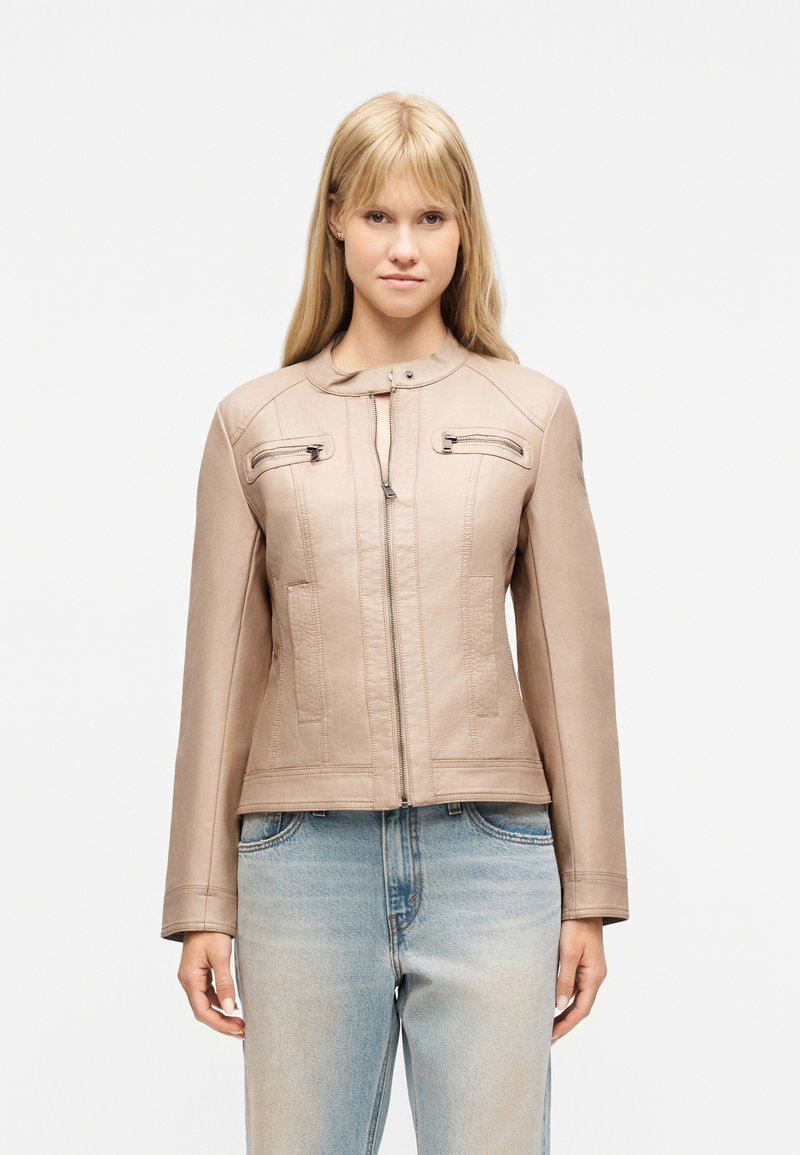Νεαρή γυναίκα που φοράει μπεζ δερμάτινο μπουφάν με εμπρόσθιο φερμουάρ και ανοιχτό μπλε τζιν, στέκεται μπροστά σε απλό λευκό φόντο.