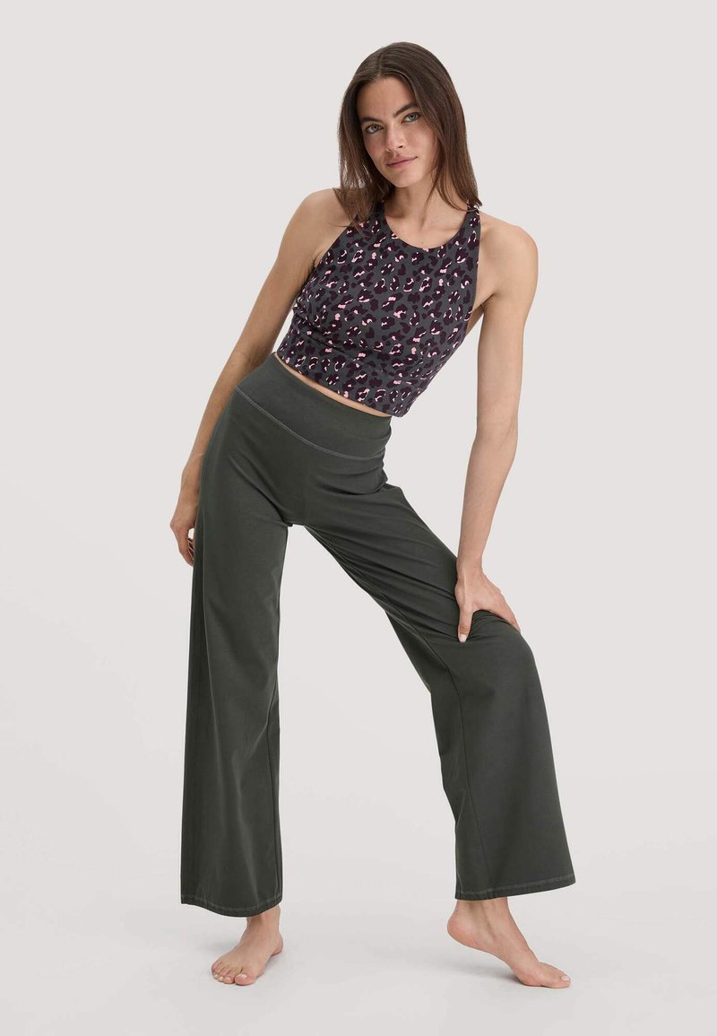 Vrouw poseert blootsvoets in een mouwloze dierenprint crop top en hoge taille wijdvallende donkere broek tegen een eenvoudige achtergrond.