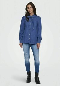 Denim srajca z gumbi ima ovratnik v obliki metuljčka, dolge rokave z nabranimi manšeti in teksturirano modro blago. Ujemata jo z raztrganimi modrimi džinsom in črnimi čevlji.