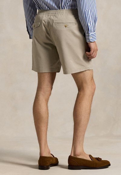 Béžové bavlněné šortky s elastickým pasem, dvě zadní kapsy se zapínáním na knoflíky, zobrazené zezadu, ve spojení s hnědými mokasínami.