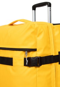 Borsa da viaggio gialla con superficie testurizzata, dotata di una cerniera nera, una tasca laterale e una tracolla nera con fibbia in plastica. Manico argento estensibile.