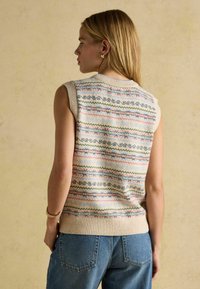 Frau mit langem, blondem Haar, die einen ärmellosen, gestrickten Pullunder mit pastellfarbenen horizontalen Mustern und Bluejeans trägt und vor einem neutralen Hintergrund steht.