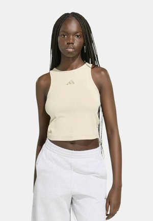 Giovane donna con lunghe trecce che indossa un top corto beige e pantaloni della tuta bianchi, in piedi con le mani nelle tasche davanti a uno sfondo semplice.