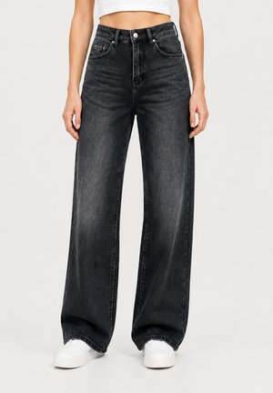 Donna che indossa jeans neri a vita alta e larghi, sneakers bianche e top corto bianco, in piedi su uno sfondo chiaro e uniforme.