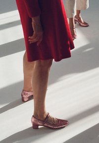 Różowe błyszczące buty Mary Jane z niskim obcasem. Buty mają zaokrąglony czubek i pasek wokół kostki. Otoczenie jest jasne i minimalistyczne.