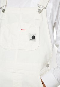 Salopette bianca in cotone con clip metalliche argentate regolabili, tasca frontale e una patch del logo grigia. Presenta dettagli di cucitura rossa sulla tasca.