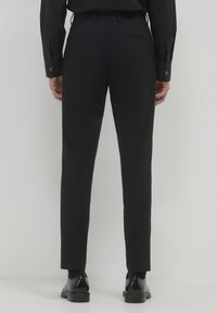Uomo in piedi di spalle, che indossa pantaloni neri slim, camicia nera con maniche cuffiate, calzini neri e scarpe eleganti nere lucide.