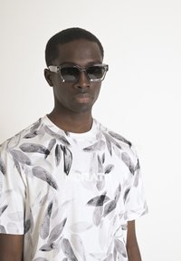 Gafas de sol negras con montura transparente, combinadas con una camiseta blanca con patrones de hojas grises y la palabra "MORATO" impresa en blanco.