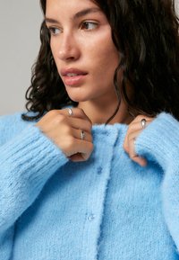 Cardigan azul claro e fofinho, com punhos canelados e fecho de botão. Apresenta uma textura suave e gola redonda, usado com anéis nos dedos.