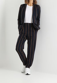 Marynarski garnitur w paski z pomarańczowymi liniami, z dopasowaną marynarką i luźnymi spodniami. Pod spodem biała koszulka. Noszony z białymi sneakersami.