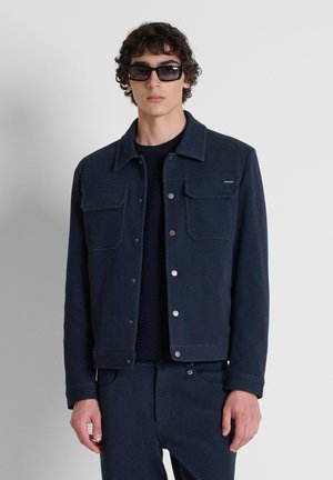 Jonge man die een donkerblauw jack met knopen en bijpassende broek draagt, met zwarte zonnebril, staand tegen een effen lichte achtergrond.