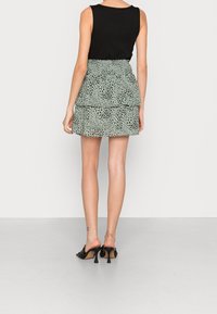 Femme portant un haut sans manches noir, une mini-jupe verte à volants avec des pois noirs, et des mules à talons noires, se tenant sur un sol uni.