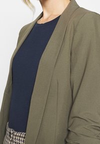 Olivově zelený blazer s volným střihem a dlouhými rukávy, přeložený přes námořnickou modrou košili. Má hladkou texturu a otevřený přední design.