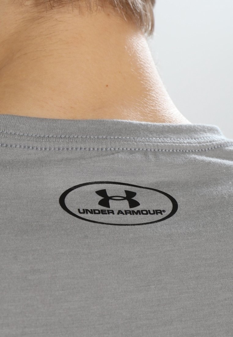 Camiseta gris de manga corta con una textura suave. Presenta un logo negro de "Under Armour" dentro de un óvalo en la parte trasera del cuello.