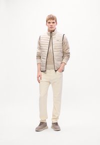 Un tânăr stă cu fața spre cameră purtând un veston bej matlasat, un pulover cu model, pantaloni crem și adidași taupe pe un fond alb.
