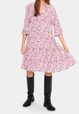 Mujer con vestido rosa floral hasta la rodilla, mangas tres cuartos y botas negras hasta la rodilla, sosteniendo el dobladillo del vestido hacia afuera.