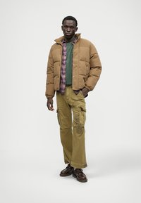 Bruine pufferjack met een groene sweater en een geruit overhemd eronder, gecombineerd met beige cargobroek met zakdetails en bruine schoenen.