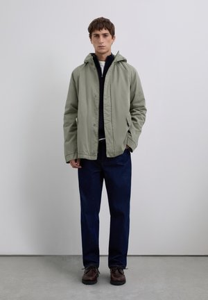 Young man standing wearing a light green hooded jacket, navy zip-up sweater, dark blue jeans, and brown lace-up shoes against a plain background.