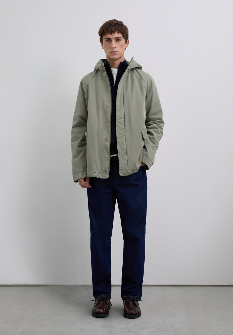 Nuori mies seisoo päällään vaaleanvihreä huppari, tummansininen vetoketjullinen villapaita, tummansiniset farkut ja ruskeat nauhakengät yksinkertaista taustaa vasten.