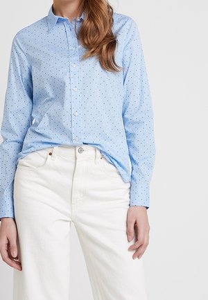 Kvinde iført en lyseblå skjorte med små sorte prikker og højtaljede hvide bukser, stående foran en ensfarvet baggrund.