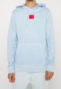 Sudadera azul claro de algodón, con un bolsillo frontal, capucha con cordón y una etiqueta de marca roja en el pecho. Textura suave.
