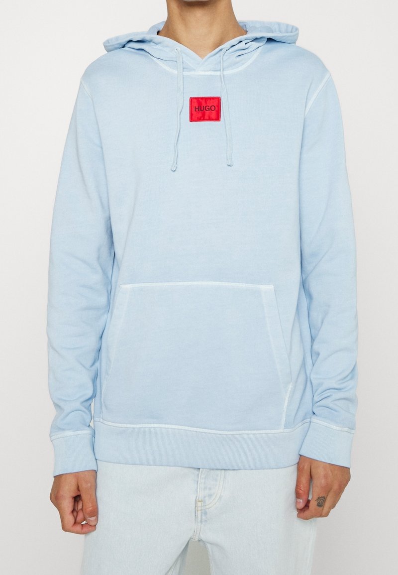 Sudadera azul claro de algodón, con un bolsillo frontal, capucha con cordón y una etiqueta de marca roja en el pecho. Textura suave.