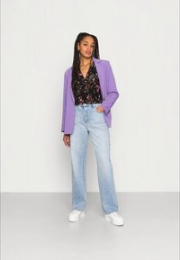Modelo vestindo um blazer lilás, uma blusa preta com padrões florais, jeans de perna larga azul claro e tênis brancos, posando sobre um fundo neutro.