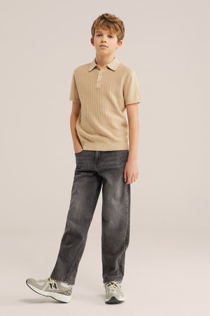 Mladi deček stoji z rokami v žepih, oblečen v bež kratkoročni pleteni polo, izbledene črne kavbojke in sive superge na enobarvnem ozadju.