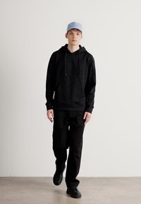 Μαύρη φόρμα με υφή, κορδόνια και μπροστινή τσέπη, συνδυασμένη με μαύρα παντελόνια και μαύρα αθλητικά παπούτσια. Το λευκό καπέλο τελειώνει την εμφάνιση.