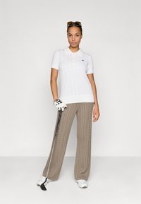 Polo bianco lavorato a maglia con maniche corte e colletto, abbinato a pantaloni a gamba larga beige con lavorazione a trecce. Tiene in mano una mazza da golf, indossando un guanto da golf.