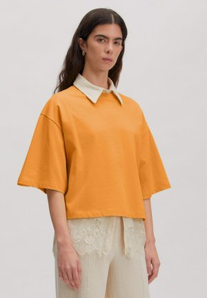 Woman with long dark hair wearing an oversized orange short-sleeve shirt over a white lace collared blouse and matching pants.
