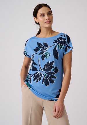 Femme aux cheveux foncés portant un haut bleu à motifs floraux et un pantalon beige, debout avec une main dans la poche, regardant légèrement vers le haut et à droite.