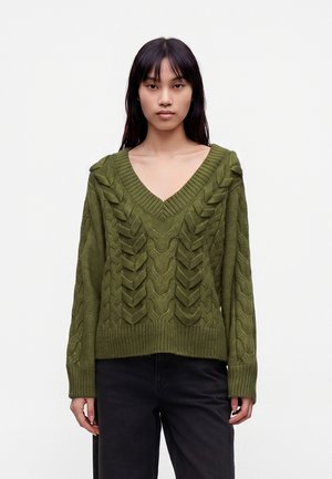 Jeune femme aux cheveux raides et foncés porte un pull vert olive à col en V en maille torsadée et un pantalon noir sur un fond clair uni.