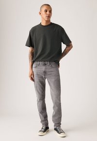 Γκρι t-shirt με λαιμόκοψη σε στυλ crew σε συνδυασμό με γκρι παντελόνι τζιν στενής εφαρμογής. Το μοντέλο φοράει ασπρόμαυρα παπούτσια τύπου high-top. Απλό, καθημερινό ντύσιμο με ελάχιστες λεπτομέρειες.