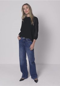 Top nero a maniche lunghe con maniche trasparenti, abbinato a jeans blu a gamba larga e ballerine nere con punta appuntita su uno sfondo neutro.