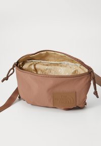 Brown fabric shoulder bag with a zippered closure, beige interior, leather logo patch, and drawstring accents on the sides.