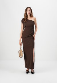 Braunes, einseitig geschultertes Kleid mit taillierter Passform und seitlichem Schlitz; kombiniert mit einer kleinen Handtasche und schwarzen, spitzen Schuhen.