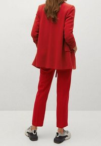 Blazer rouge ajusté et pantalon assorti en tissu lisse. Stylé avec des baskets sportives grises. Vue arrière mettant en valeur le design ajusté.