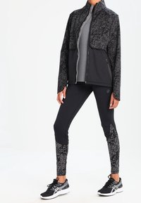 Chaqueta deportiva negra con patrón geométrico, camiseta gris con cremallera y leggings negros con diseños a juego. Zapatillas negras completan el conjunto.