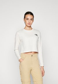 Maglietta a maniche lunghe di colore bianco sporco con logo nero piccolo sul lato sinistro del petto. Indossata con pantaloni cargo beige chiaro.