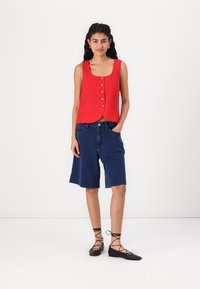 Red sleeveless top with round neckline and front button details, paired with navy denim shorts. Model wears black lace-up flats.