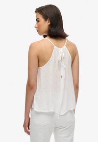 Witte mouwloze top met verstelbare strik aan de achterkant, lichtgewicht stof, losse pasvorm, afgeronde zoom en subtiele textuurdetails.