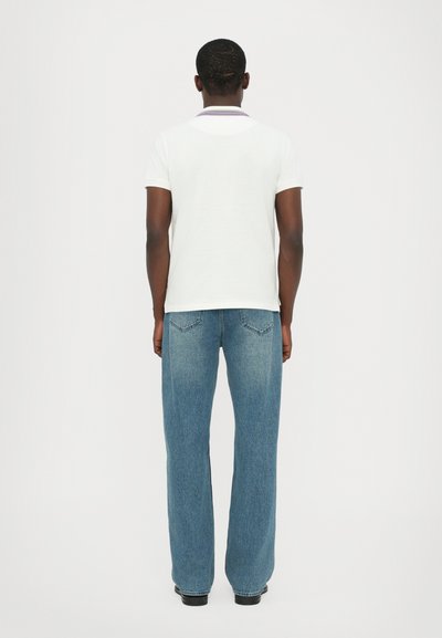 Balta polo krekls ar purpura apkakli, apvienots ar gaiši zilu platu biksu, rādīts no aizmugures uz vienkārša fona.