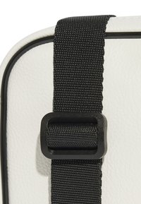 Angolo di una borsa in pelle bianca con una tracolla in nylon tessuto nero caratterizzata da una fibbia di plastica rettangolare. Finitura liscia con cuciture sottili.