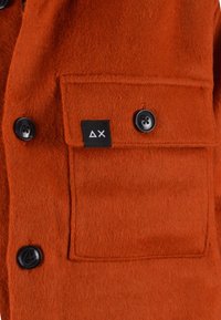 Giacca di lana arancione con tasca a patch e bottoni neri. Il tessuto ha una consistenza morbida con un'etichetta che mostra un logo geometrico.