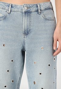 Lys blå denim jeans med grommetdetaljer langs bena, en tettsittende midje og klassisk femlomme-design. Teksturert, middels tykkelse.