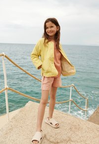 Gul quiltet jakke, rosa t-skjorte, lyserosa shorts og beige sandaler. Bakgrunnen viser steinete kystlinje og hav.