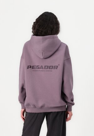 Person med krøllet hår iført en løs mauve hoodie med "PEGADOR"-logo på bagsiden og sorte bukser, stående mod en ensfarvet hvid baggrund.